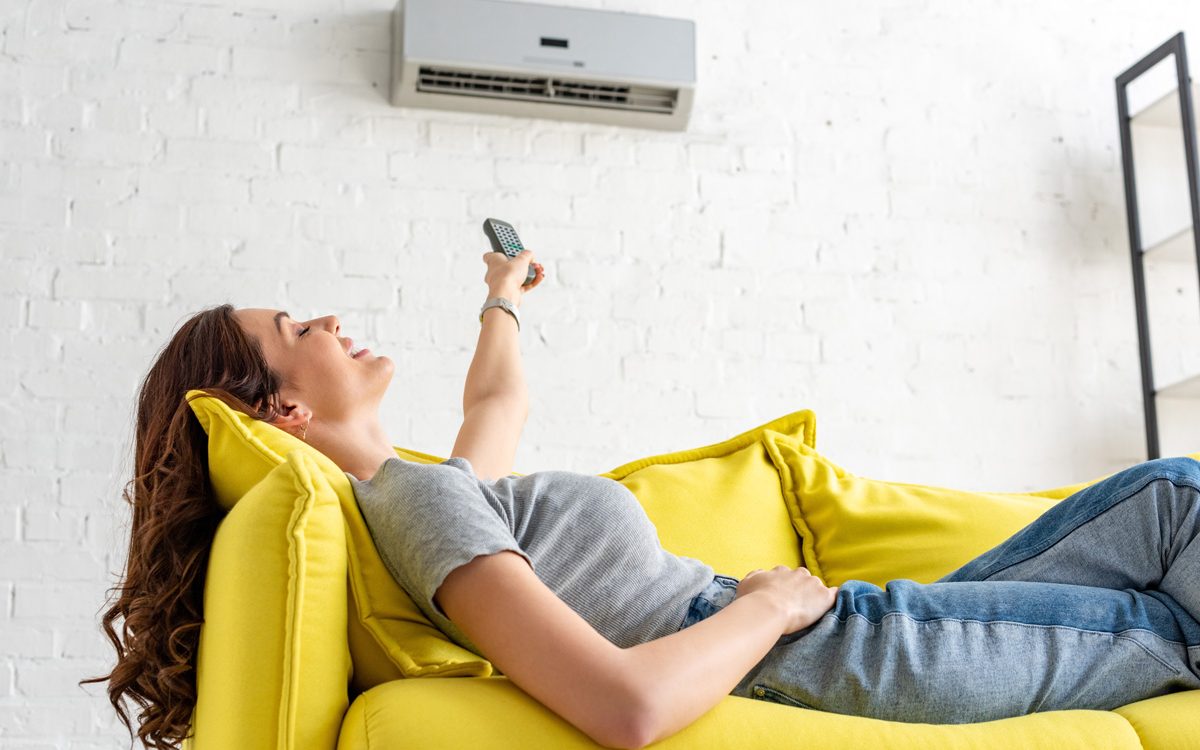 A person laying on a yellow couch turning on a wall AC unit with a remote in North Fort Myers.